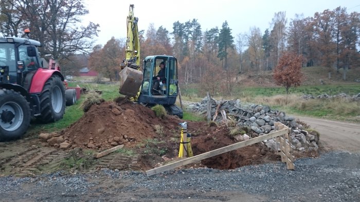 Grävmaskin gräver stora hål för gjutning av stolpar till grindar på en lerig byggarbetsplats, med traktor i bakgrunden.