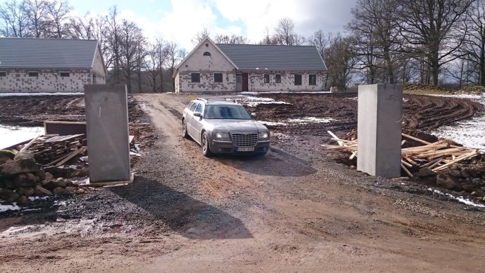 Bil parkerad på grusväg framför ett garage under konstruktion med tunga stolpar på sidorna och en del snö kvar på marken.