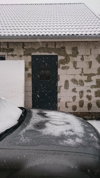 Delvis snötäckt bil framför ett ofärdigt garage med snö på taket och snöfall.