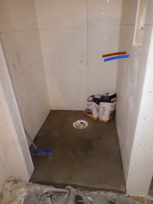 Ett delvis flytspacklat badrumsgolv med golvbrunn, omgivet av väggar med kakel och avstängningslister synliga.