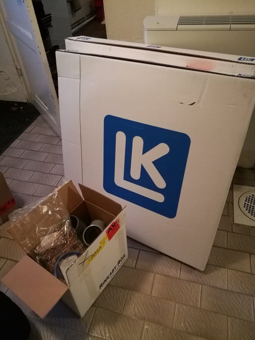 Stor vit LK förpackning och öppen kartong med VVS-delar inklusive rör och kopplingar på ett kaklat golv.