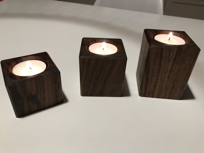 Tre handgjorda värmeljushållare i valnöt, oljade, med tända ljus på vit yta.