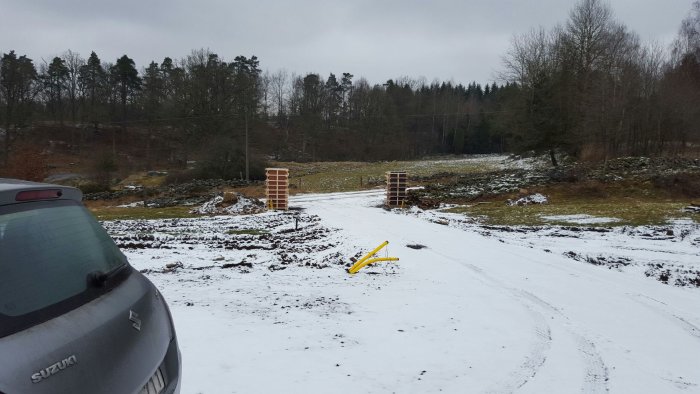 Två betongpelare för grindar på en uppfart täckt av snö, med armeringsjärn och gul byggutrustning.