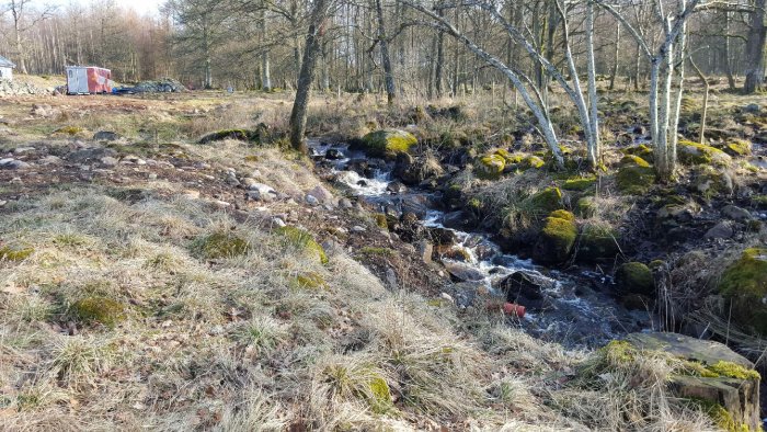 En bäck omgiven av mossbeklädda stenar och vissna grästuvor rinner genom en tomt med lite snö och en byggarbetsplats i bakgrunden.