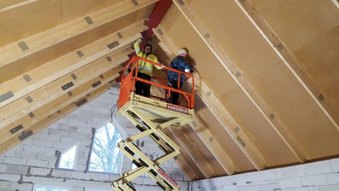 Två personer i arbetskläder står på en skylift inuti en byggnad under konstruktion, arbetar med taket.