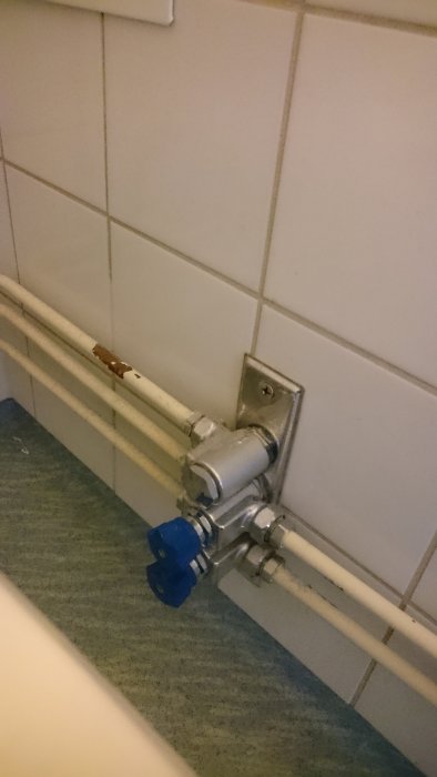 Rörinstallation med en blå handtag kran monterad på vit kakelvägg.