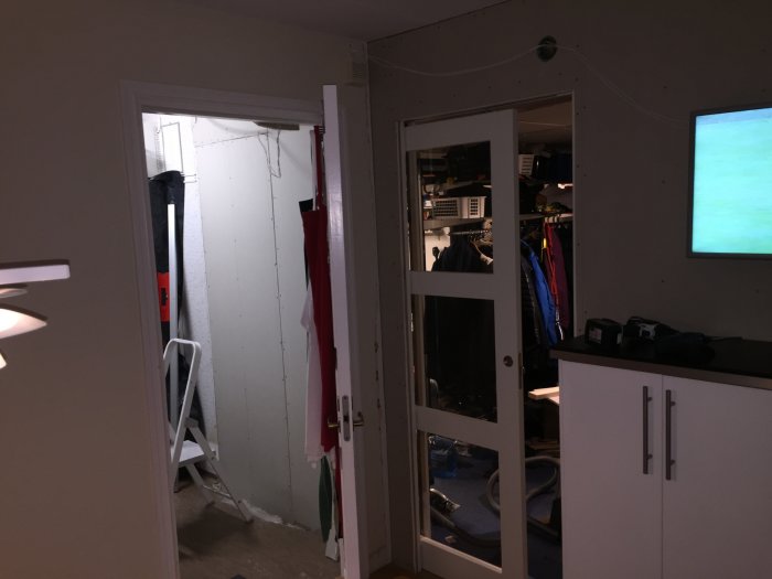 Interiör av ett hem under renovering, visar gipsskivor längs en vägg med en dörröppning och en vit stege, samt delar av ett kök.