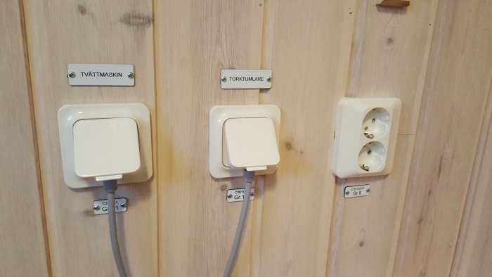 Eluttag och vägguttag märkta med skyltar 'TVÄTTMASKIN' och 'TORKTUMMARE' på en trävägg.