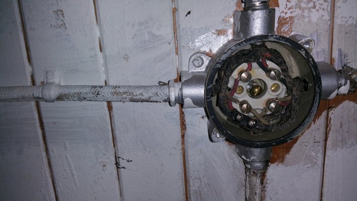 Öppnad elektrisk dosa med exponerade ledningar och anslutningsterminaler på en vägg med vit färg.