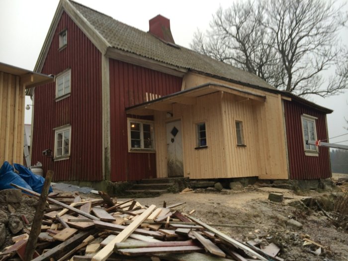 1700-tals byggnad under renovering med nytt träväggssektion och röra av byggmaterial framför huset.