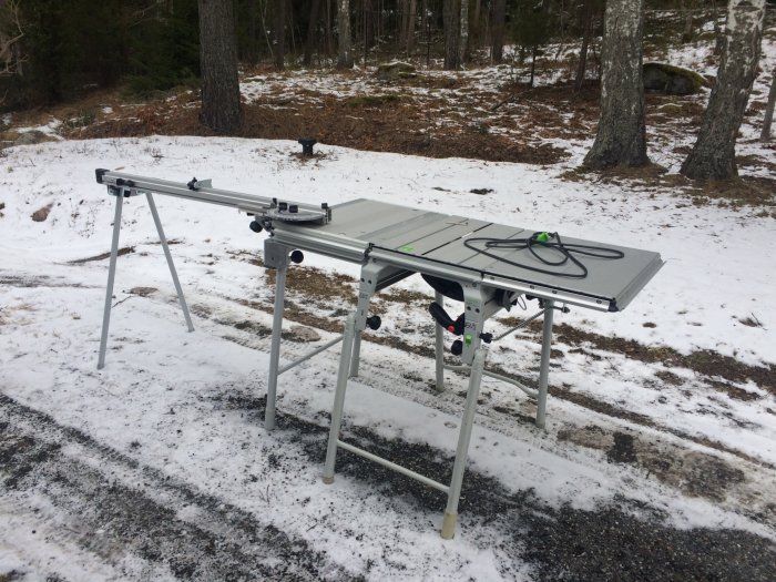 En hopfällbar sågbord står utomhus på en snöfläckig mark med tillbehör för sågning fäst vid bordet.