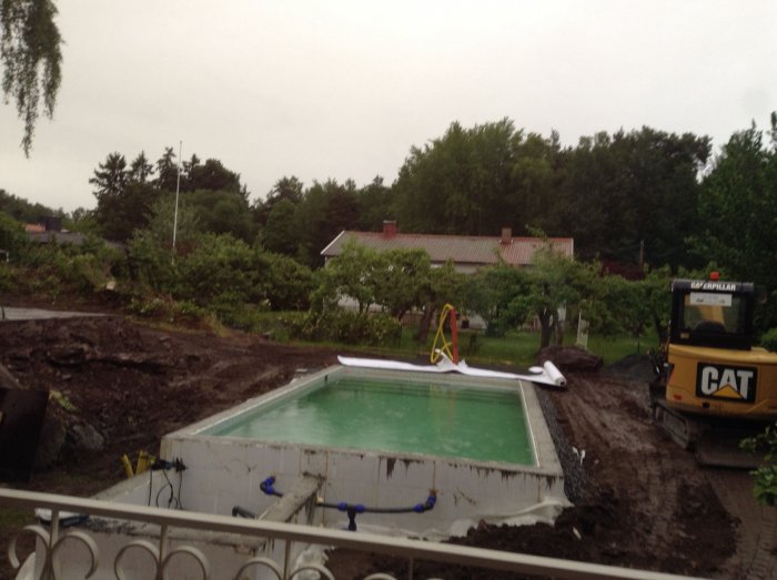 En pool under konstruktion med grävmaskin och hopfälld rutschkana i en trädgård.