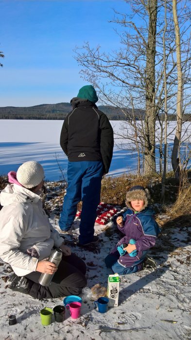 Person och barn vid fruset vatten med termos och muggar i soligt väder.