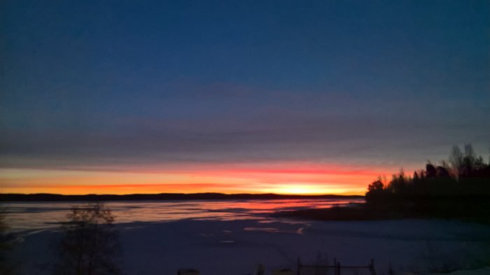 Solnedgång över ett fruset landskap med silhuetter av träd och byggnader.
