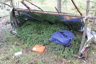 Primitiv bivacklägerplats i skogen med grenar, presenning och sovsäck.