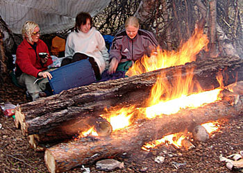 Tre personer sitter nära en stor brasa utomhus med campingutrustning.