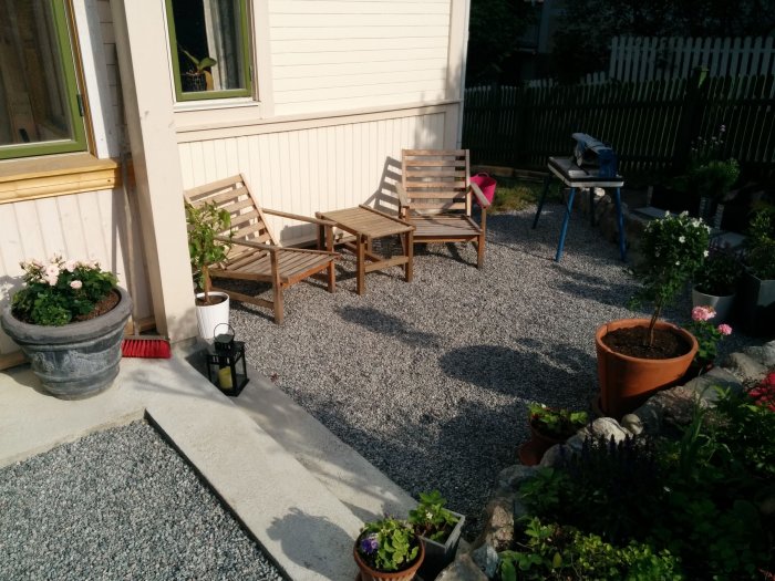 Grusad uteplats med trämöbler och krukväxter vid en vit husvägg i soligt väder.
