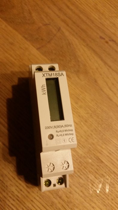 Energimätare med digital display och tekniska specifikationer på framsidan, placerad på ett träbord.