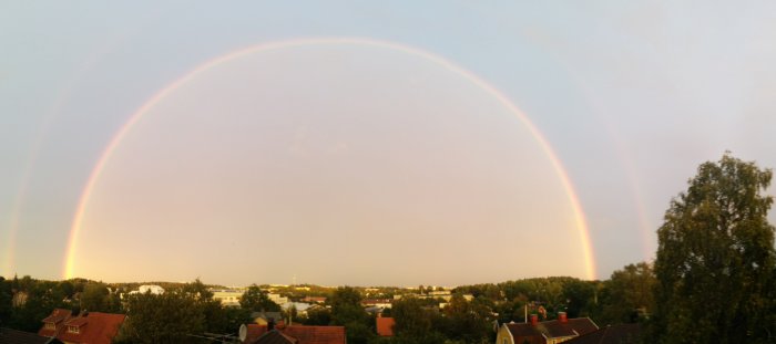 Panoramavy från ett fönster med en stor regnbåge som bildar en båge över en förort.