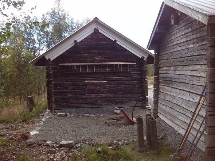 Timrad stuga med tillbyggnad under konstruktion, synlig regelstomme och fundament.