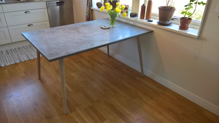 Köksbord med ben i ask och skiva i expanderbetong intill en vit vägg och fönster med blommor.