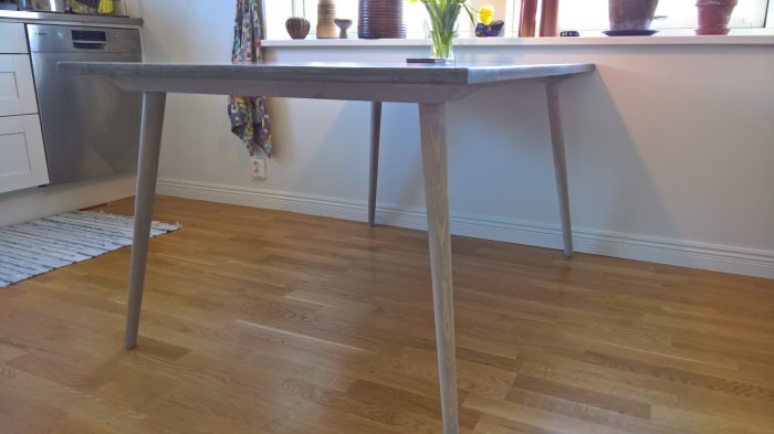 Köksbord med ben i ask behandlade med grå tråsåpa och skiva i expanderbetong, intill fönster och vägg.