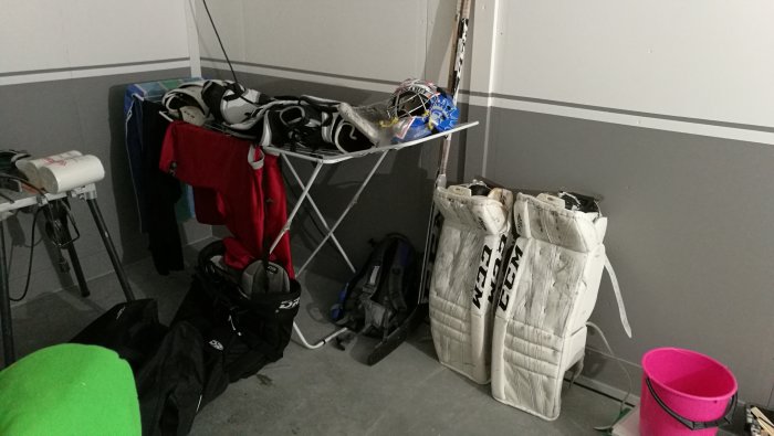 Ishockeyutrustning förvaras i ett garage, inklusive målvaktsbenskydd och hjälm på en torkställning.