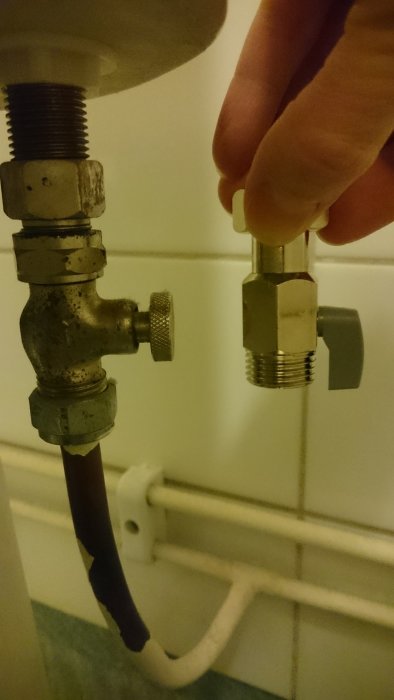 Hand som håller en gammal mutter nära rörkopplingar och avstängningsventil i badrum.