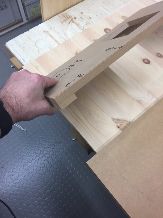 Hand håller trämall för komplicerade hål, med markerade referenslinjer, över en träbit.