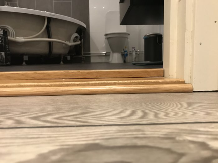 Närbild på en uppstickande dörrlist vid badrumsdörr med sprucken fog synlig, dörr och toalett i bakgrunden.