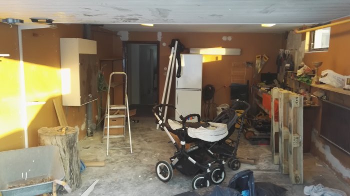 Oorganiserad garageinteriör med diverse föremål och byggmaterial utspridda på golvet samt barnvagn.