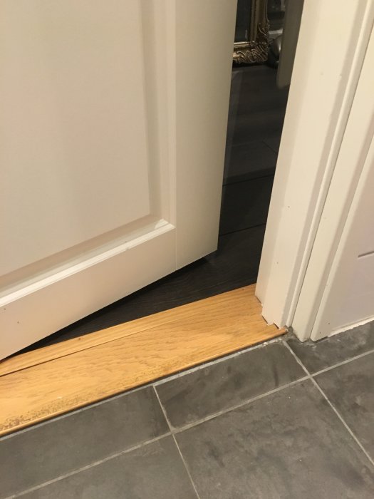 Golvet vid en dörröppning visar en trätröskel bredvid grå klinkerplattor med en synlig glipa.
