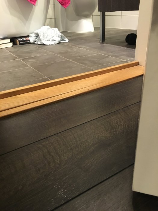 Tröskel mellan grått klinkergolv och mörkt trägolv med golvlist och dörrpost synliga.