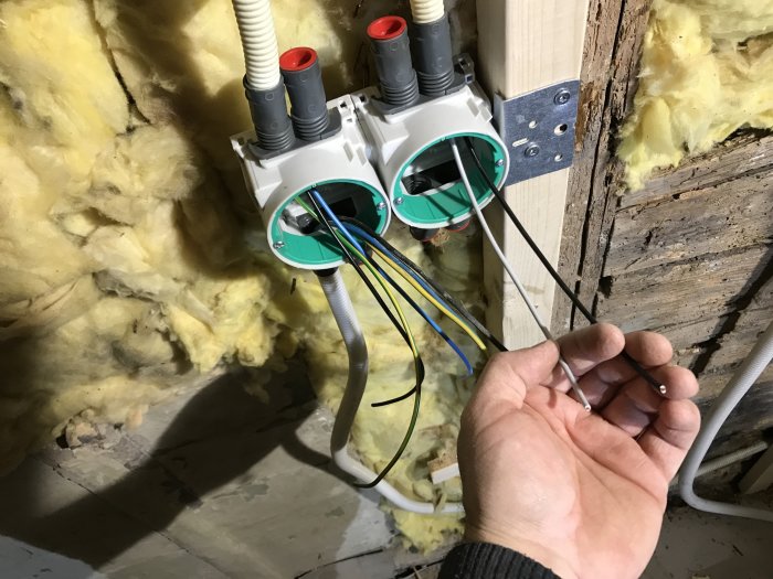 Hand som håller elektriska kablar framför dosor monterade på regel med isolering i bakgrunden.