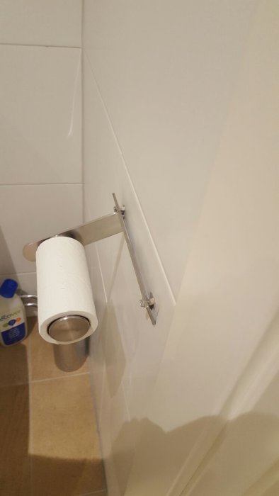Toalettpappershållare monterad på kakelvägg, med synliga skruvar och potentiellt behov av tätning eller lim.