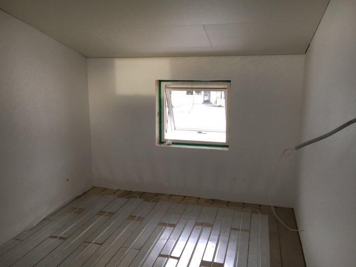 Rum under renovering med nylagda oanslutna golvvärmeslingor och oputsat fönster.