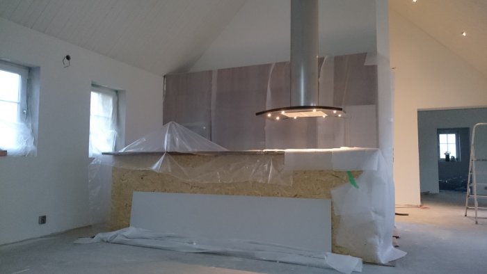 Köksrenovering med en oskyddad köksö under en hängande fläkt, omgiven av byggmaterial och plastövertäckt inredning.