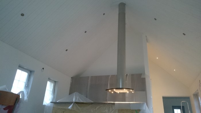 Köksrenovering med en modern köksfläkt i rostfritt stål som monteras ovanför en skyddad köksö, inomhusarbete pågår.