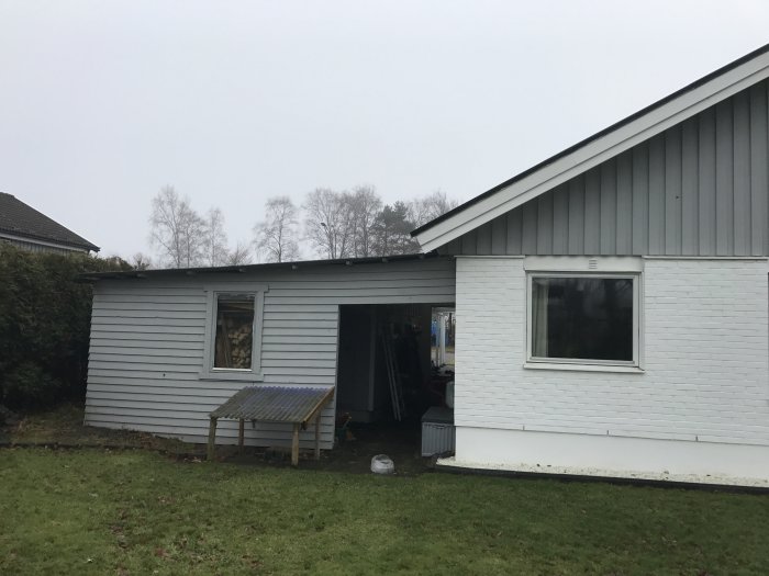 Ett hus med vit fasad och en carport, grå sidobyggnad med öppen dörr och vedstaplar synliga.