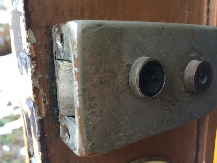 Gammalt lås fastskruvat på en ytterdörr med två synliga skruvhål, ser ut att vara svårt att montera bort.