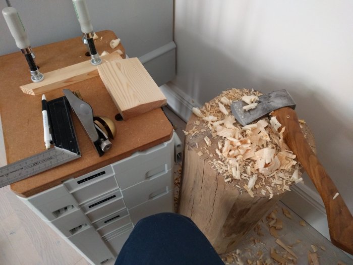 Hantverk med yxa, hyvel och skurklossar omgivna av träspån på arbetsbänk.