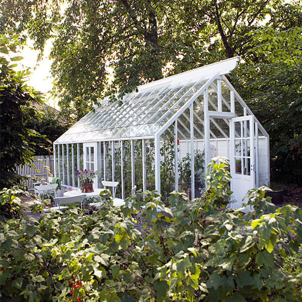 Stort, ljust växthus med vitmålade limträbalkar omgärdat av grönska och en bänk framför.