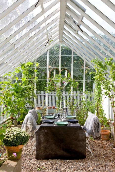 Inviting greenhouse interior with a large wooden beam, lush plants, a dining table set for two, and a white picket fence in the background.