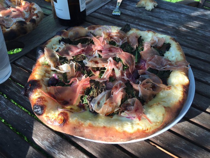 Hembakad pizza med prosciutto och grönsaker på ett träbord utomhus.