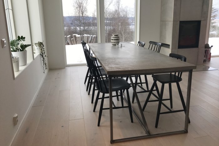 Betongbord med tvärgående stag och svarta stolar i ett ljust rum med utsikt över ett snöigt landskap.