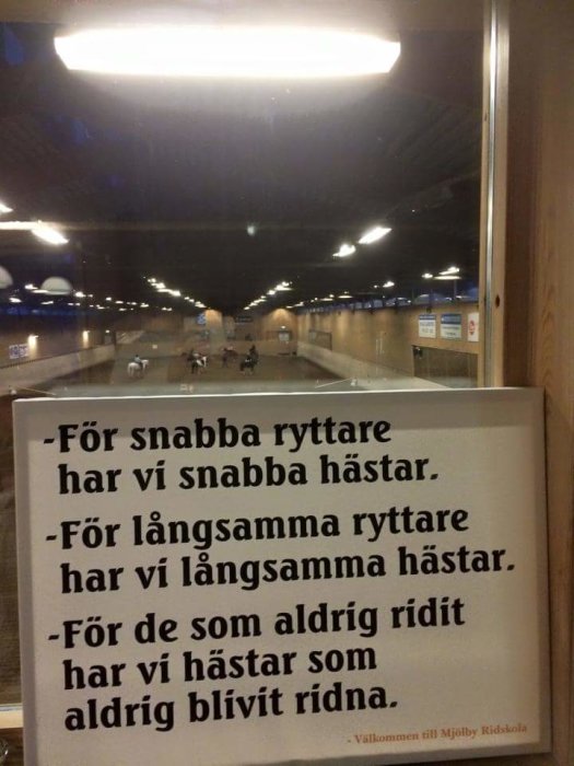 Rid-skylt med text som skämtsamt anpassar typ av häst efter ryttarens erfarenhet, reflekterad i ett fönster som visar en ridbana.