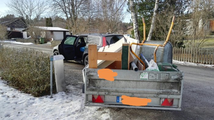 Full container för byggavfall med träbitar, metall och verktyg, parkerad vid en snötäckt väg.