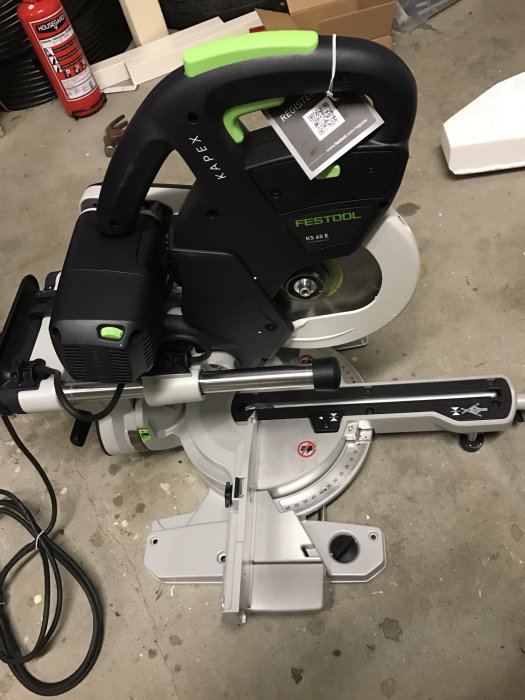 Ny kap-/gersåg av märket Festool KAPEX KS 60 E på ett garagegolv med brandsläckare i bakgrunden.