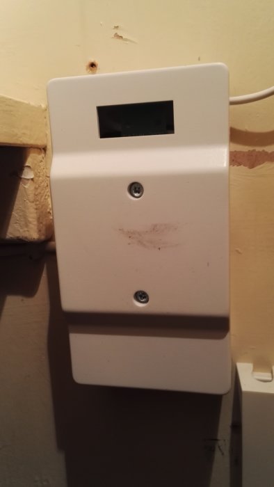 Vit termostat på vägg med display och två skruvar, smutsfläckar synliga runtomkring.