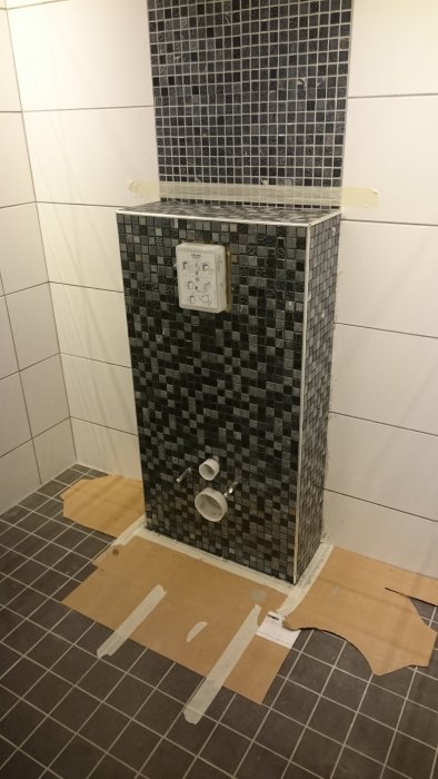 Badrum under renovering med svart mosaik, synligt avlopp och inbyggnadstoalett i vägg, omgivet av skyddspapp.